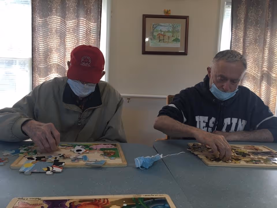 Two elderly men sitting at a table working on wooden puzzles. Both are wearing face masks, one man is wearing a red cap and a beige jacket, the other is wearing a dark sweatshirt. There are windows with patterned curtains behind them and a framed picture on the wall.