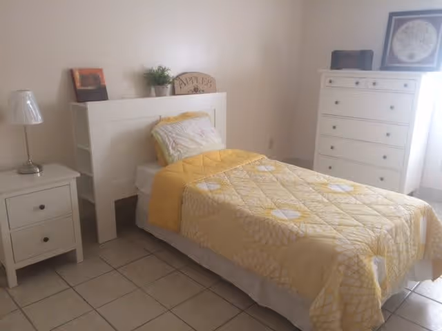 A simple bedroom with a single bed covered in a yellow and white quilt, a white headboard with shelves, a nightstand with a lamp, and a white dresser with a framed picture on top. The room has tiled flooring and light-colored walls.