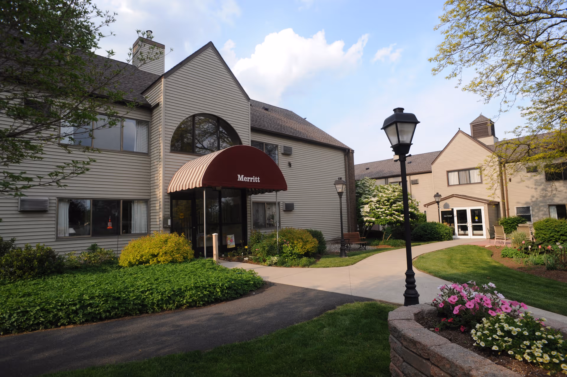 Exterior view of a senior living facility building with beige siding and a maroon awning labeled 'Merritt' over the entrance. The building is surrounded by well-maintained landscaping including green bushes, a flower bed with pink and white flowers, and a paved walkway. There are black lamp posts and benches along the walkway under a partly cloudy sky.