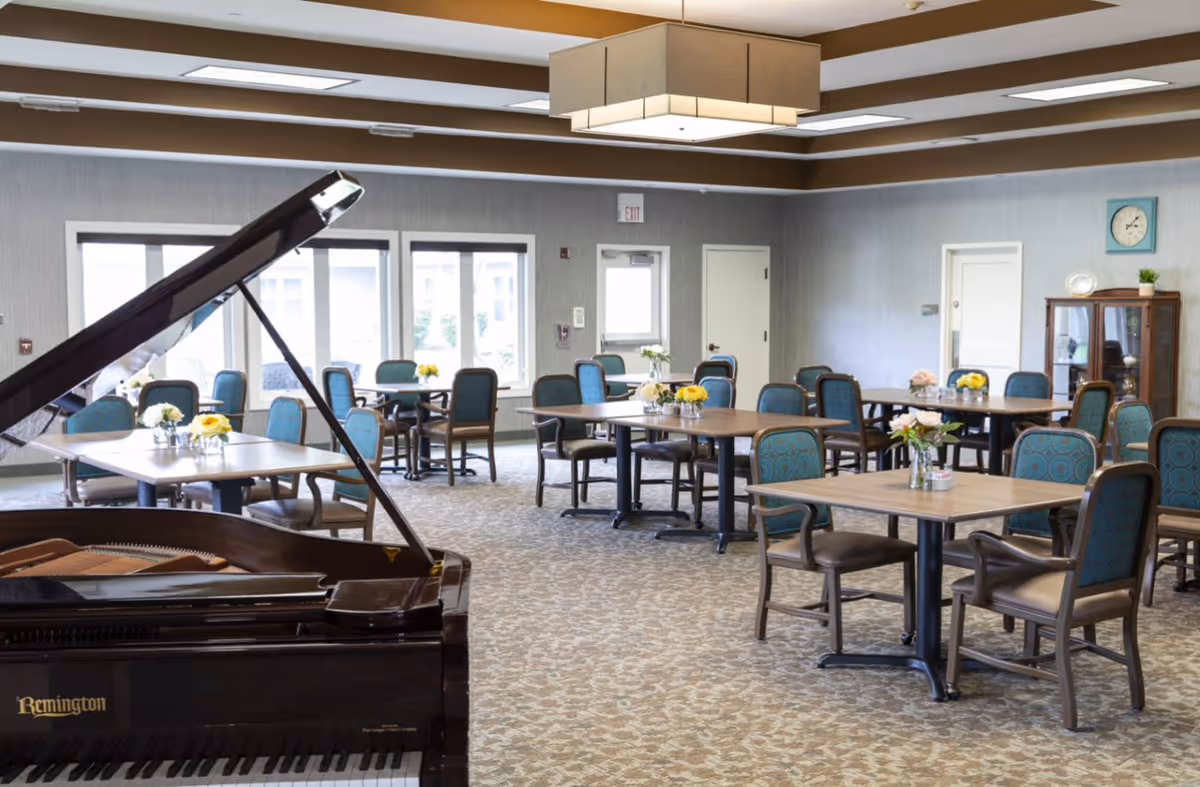 Spacious dining/activity room with multiple tables and chairs, floral centerpieces, and a grand piano in the foreground.