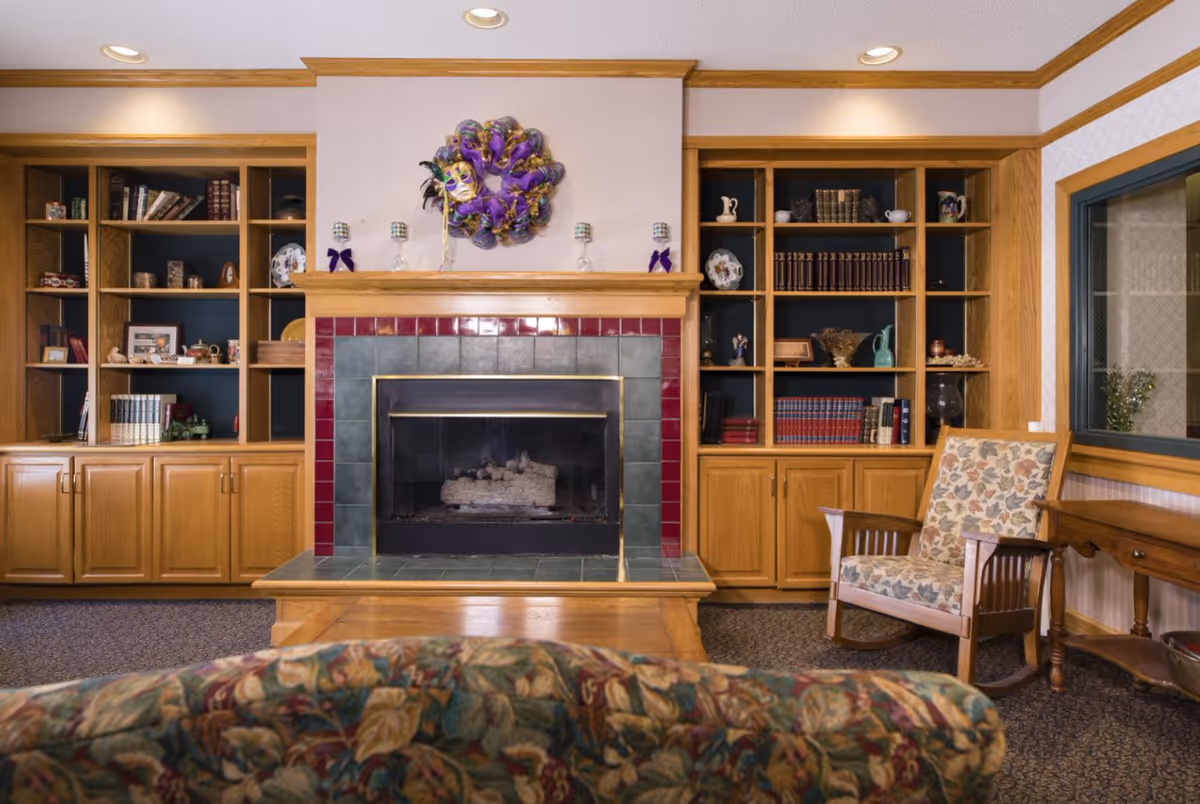 Interior common room with a tiled fireplace centered between wooden bookshelves, a floral-upholstered couch and armchair.