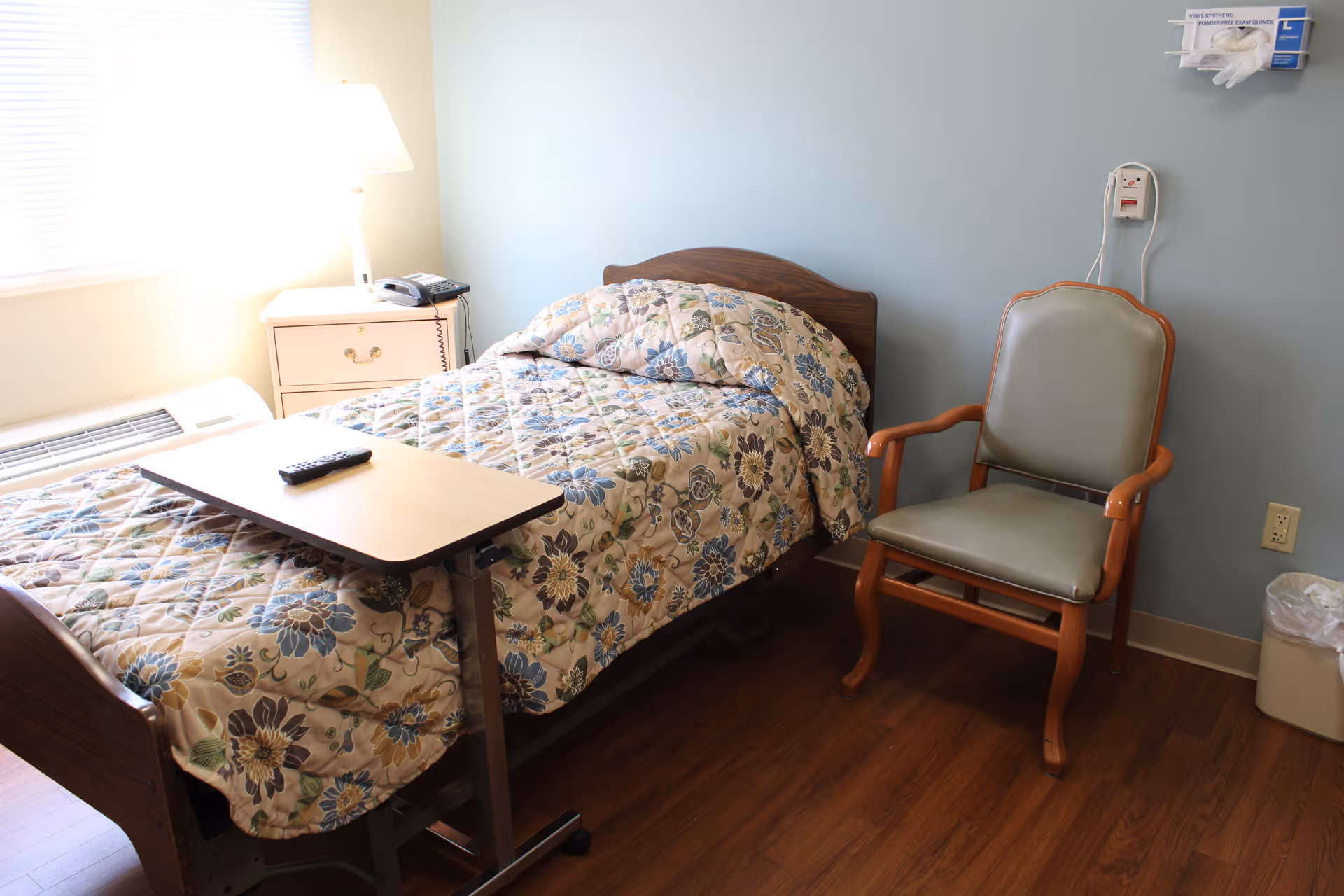 A small bedroom in a senior living facility with a single bed covered in a floral quilt, a wooden bedside table with a lamp and telephone, a remote control on an overbed table, a padded wooden chair, and a wall-mounted box of disposable gloves.