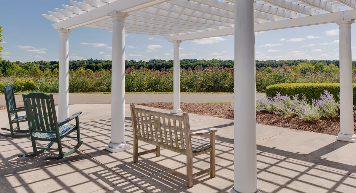 Outdoor seating area with a wooden bench and two green rocking chairs under a white pergola. The area overlooks a scenic view of greenery and a clear blue sky with scattered clouds.