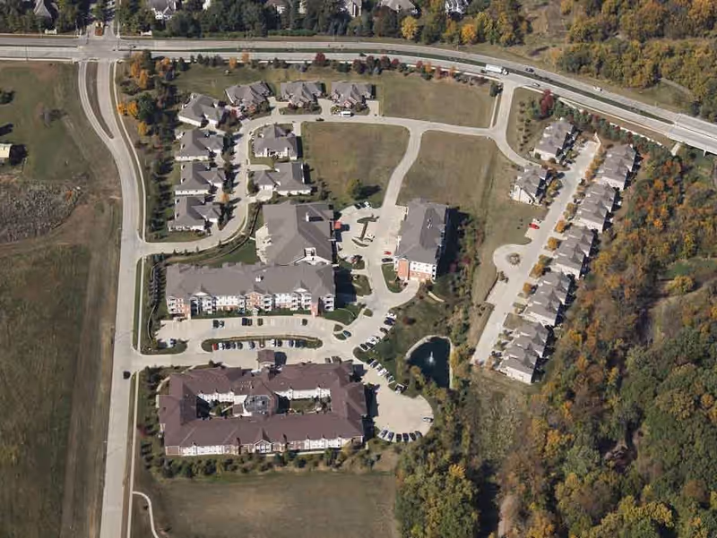 Aerial view of Silvercrest at Woodlands Creek senior living facility showing multiple residential buildings, parking areas, roads, and surrounding greenery with trees and open fields.