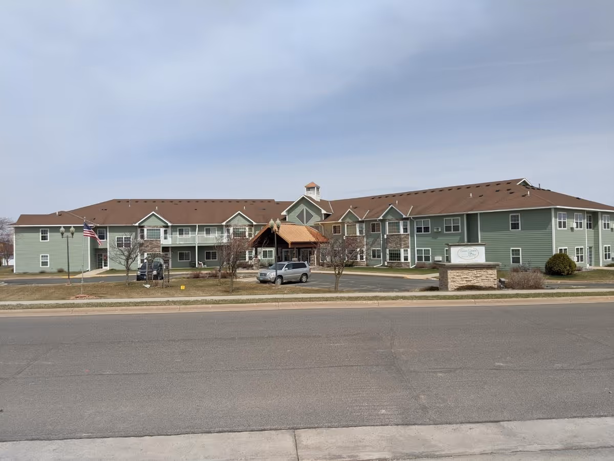 Exterior view of a two-story senior living facility building with green siding and a brown roof. There is a covered entrance with a peaked roof in the center, a few cars parked in front, an American flag on a pole to the left, and a sign on a stone base near the entrance. The sky is clear with some light clouds.