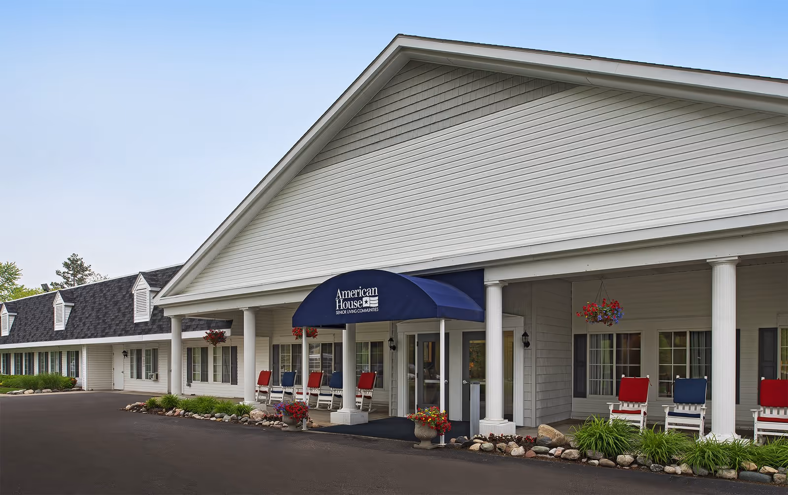 Exterior front view of American House Dearborn Heights senior living community building with a covered entrance, white columns, and a blue awning displaying the facility name. There are red and blue rocking chairs on the porch and hanging flower baskets.
