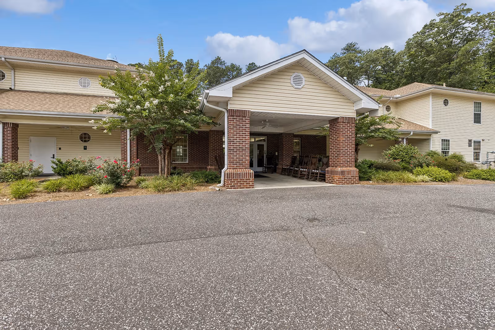 Covered entrance with brick columns and rocking chairs at the front of a two-story senior living building surrounded by landscaping.