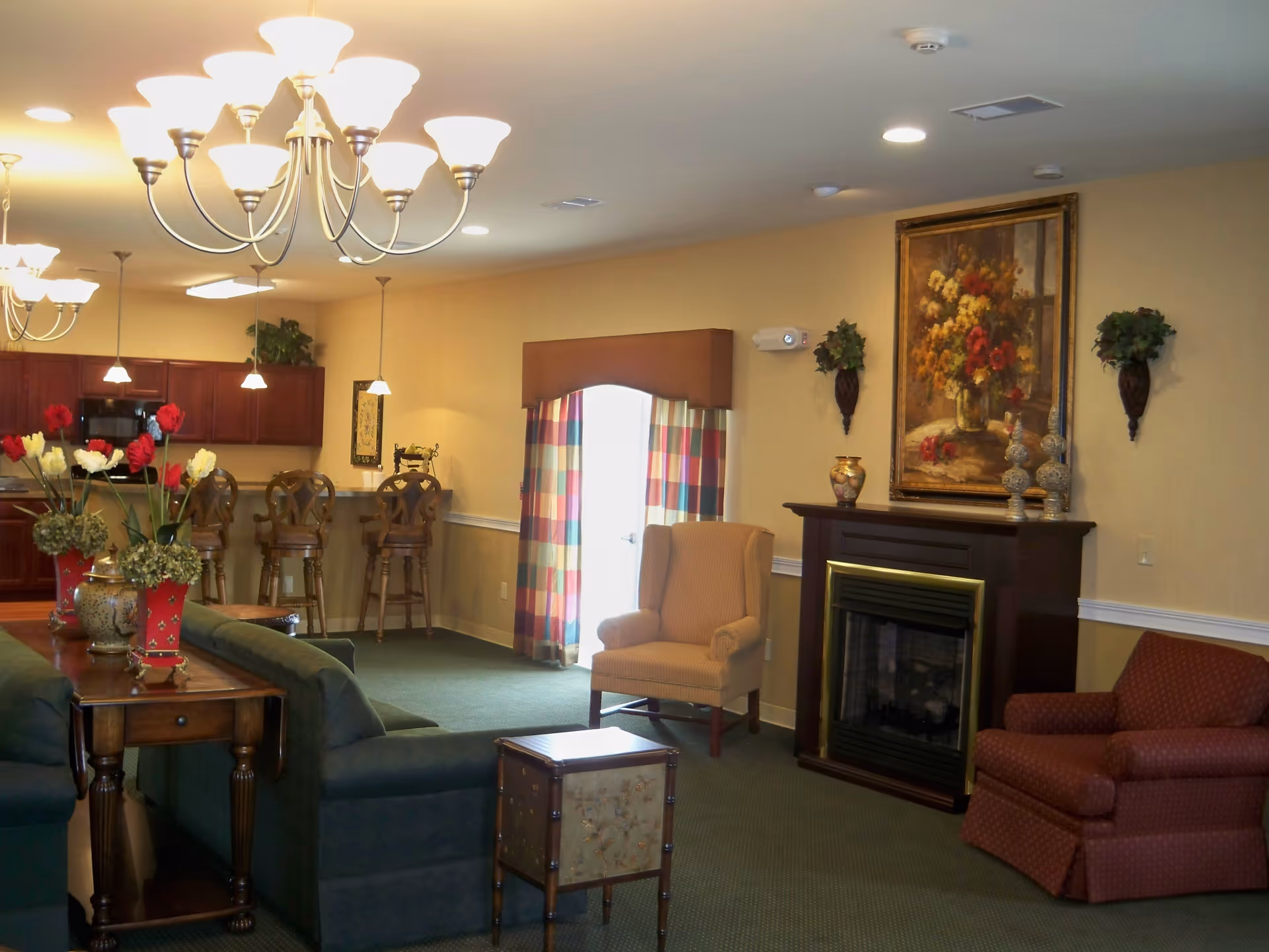 A cozy living room area in The Willows at Bellevue featuring green sofas, a wooden side table with flower vases, two armchairs near a dark wood fireplace with decorative items and a large floral painting above it. In the background, there is a kitchen area with wooden cabinets, a counter with bar stools, and pendant lighting. The room is warmly lit with chandeliers and recessed ceiling lights, and a door with colorful curtains is visible on the far wall.