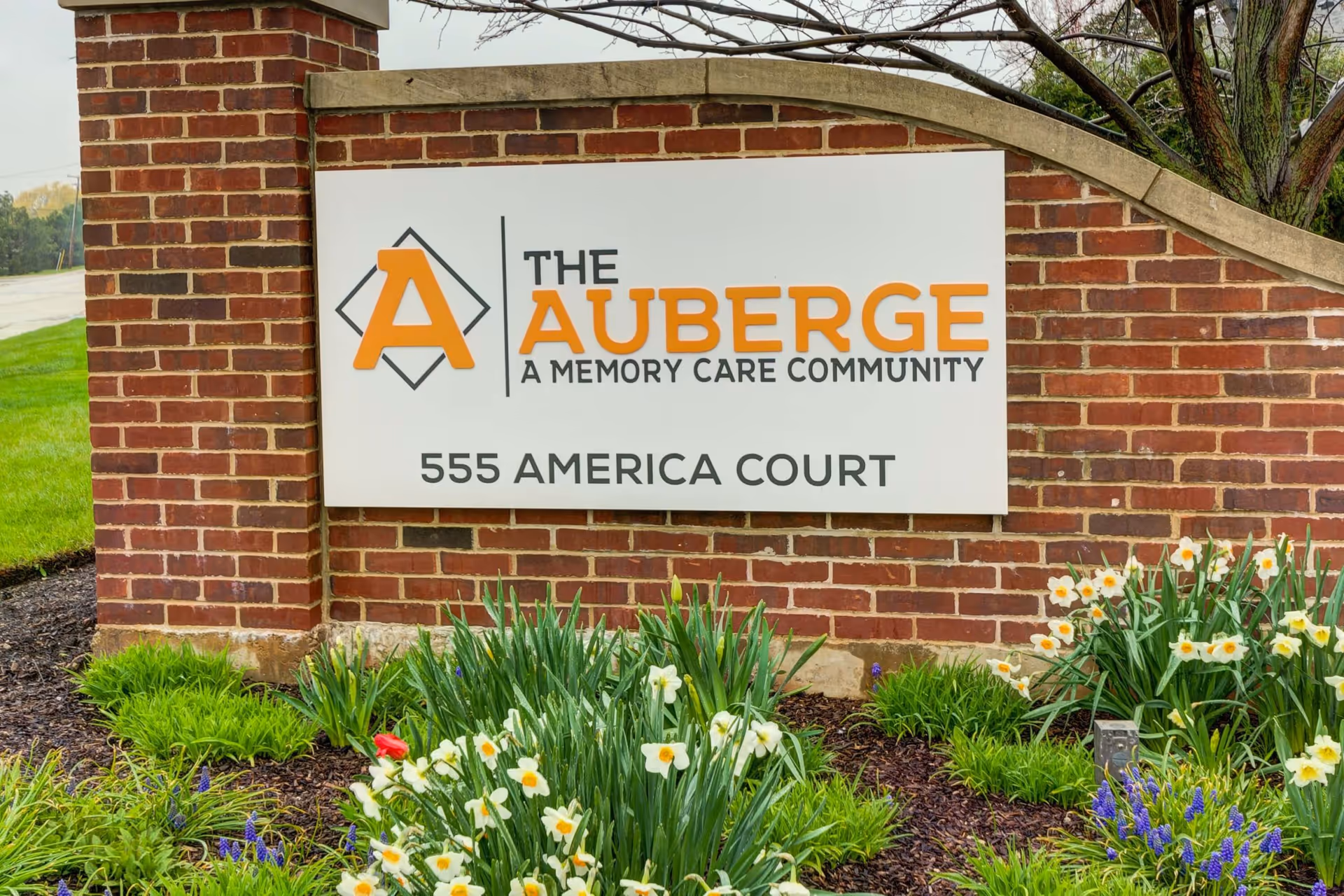 Brick entrance sign reading "The Auberge A Memory Care Community 555 America Court" surrounded by spring flowers and landscaping.