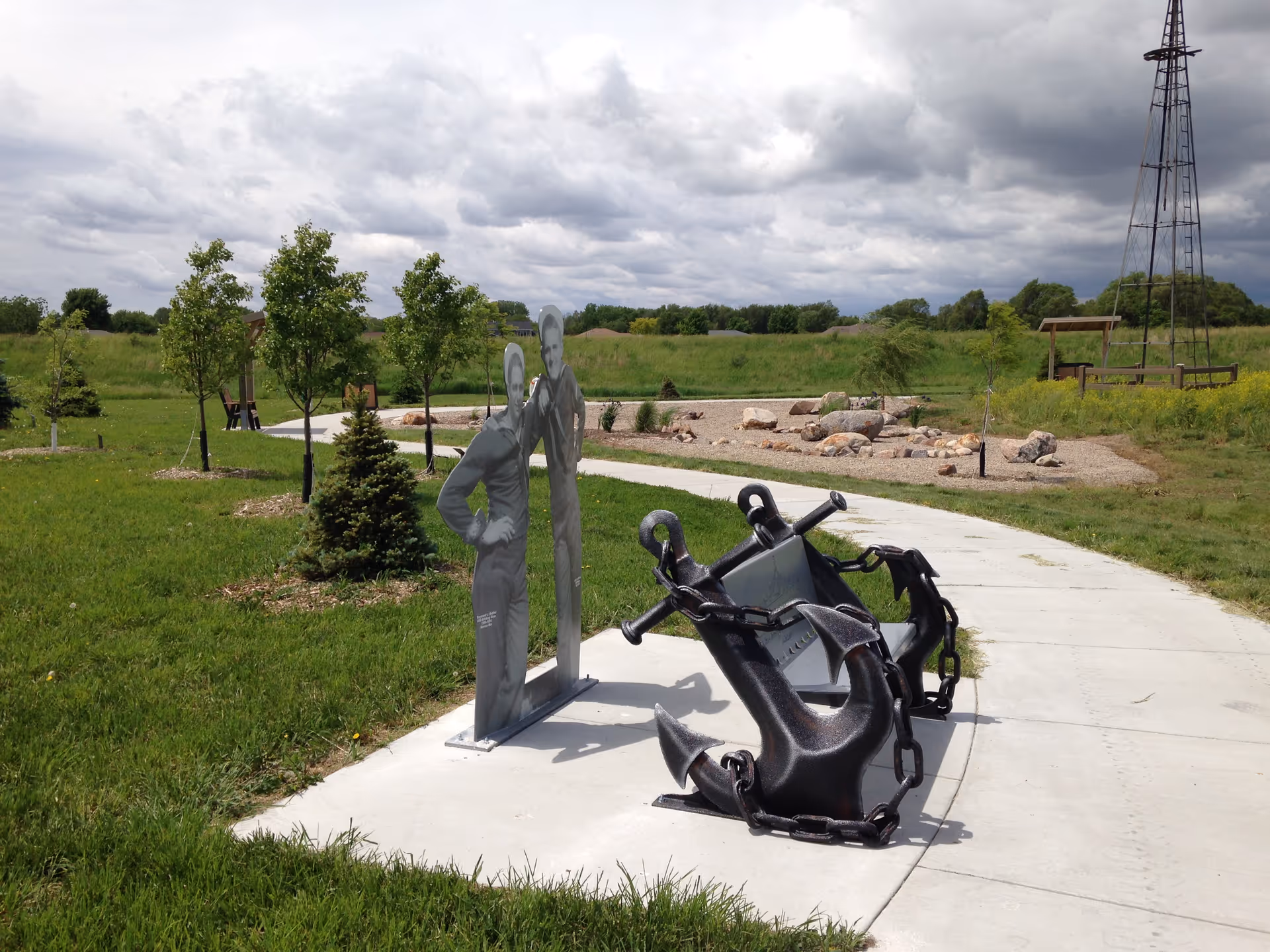 Outdoor pathway in a grassy area with small trees and shrubs. Along the path, there is a large black anchor sculpture and a metal cutout sculpture of two people. In the background, there is a windmill structure and a wooden bench under a small shelter. The sky is cloudy.