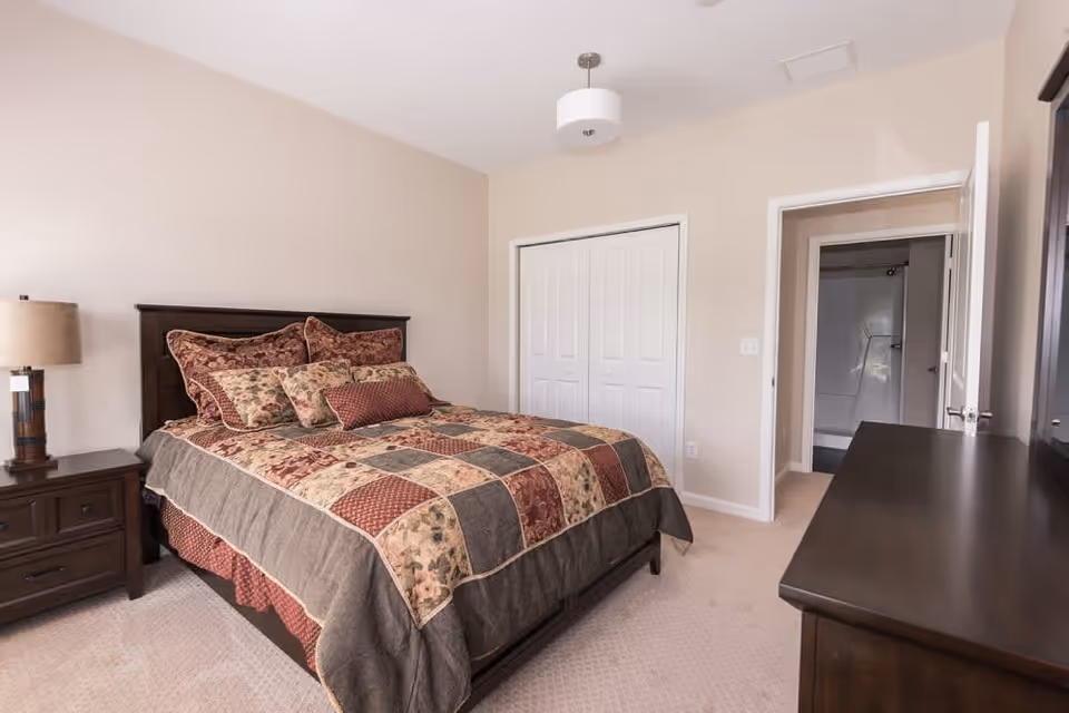 A bedroom with a bed featuring a patchwork quilt and multiple pillows. There is a wooden nightstand with a lamp on the left side of the bed. A dresser is visible on the right side of the image. The room has beige walls, a white closet with double doors, and an open door leading to a bathroom with a bathtub and shower.