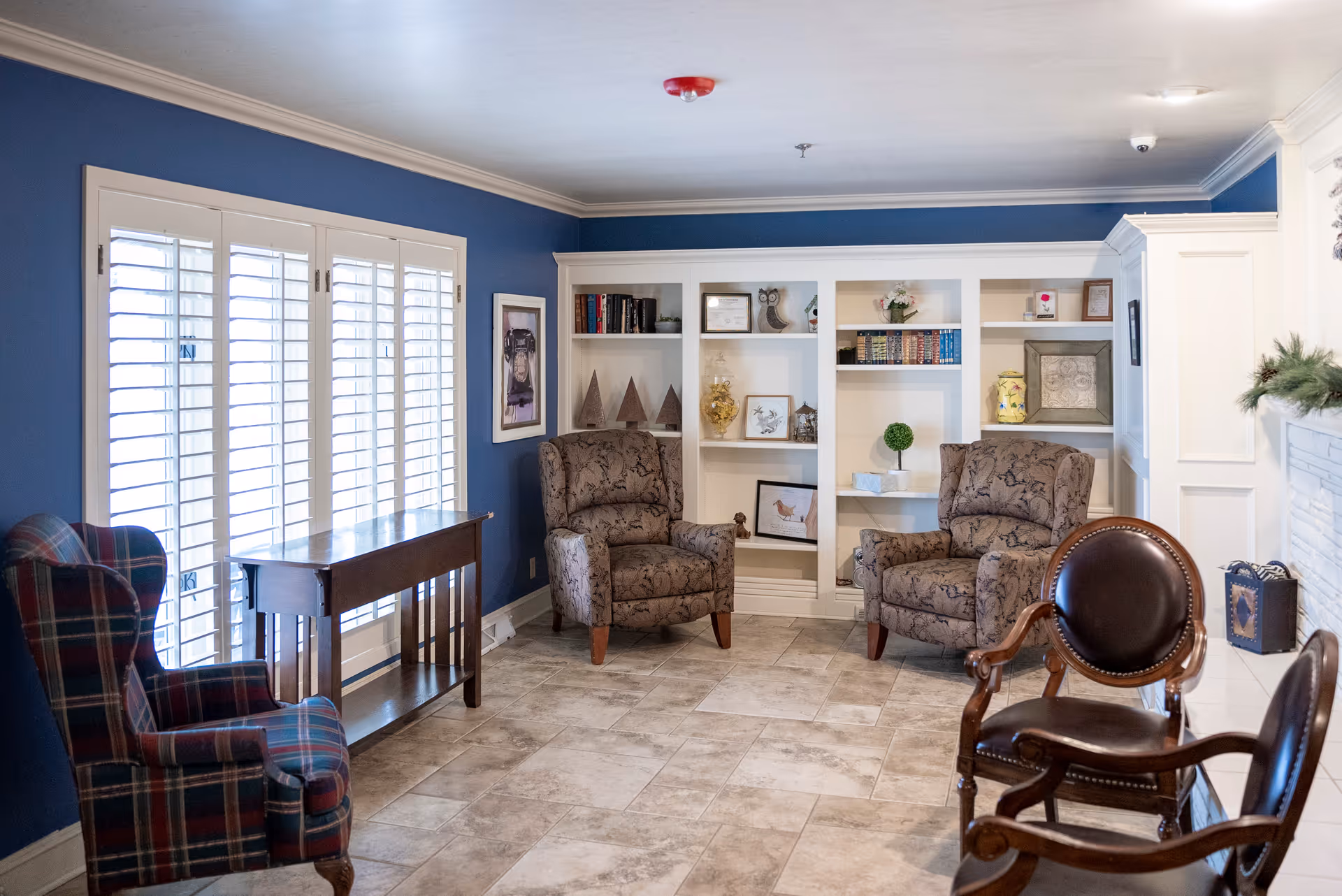 Bright communal sitting room with blue walls, white built-in bookshelves, plantation-shuttered windows, and several upholstered armchairs.