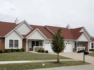 Single-story brick and white siding residential building with red gabled roofs, attached garages, small porches, and a front lawn with a young tree.