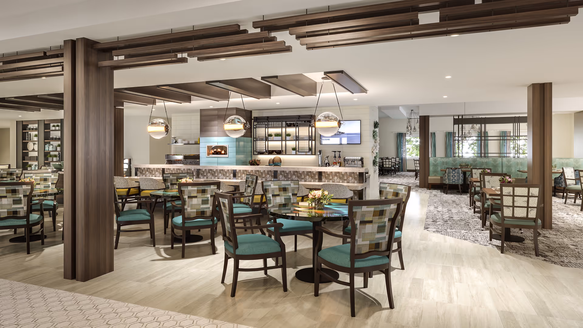 A spacious and modern dining area with multiple round tables and chairs featuring teal cushions and patterned backs. The room has wooden ceiling accents, pendant lights, and a counter area with a built-in oven and shelves with decorative items. Large windows with curtains allow natural light to brighten the space.