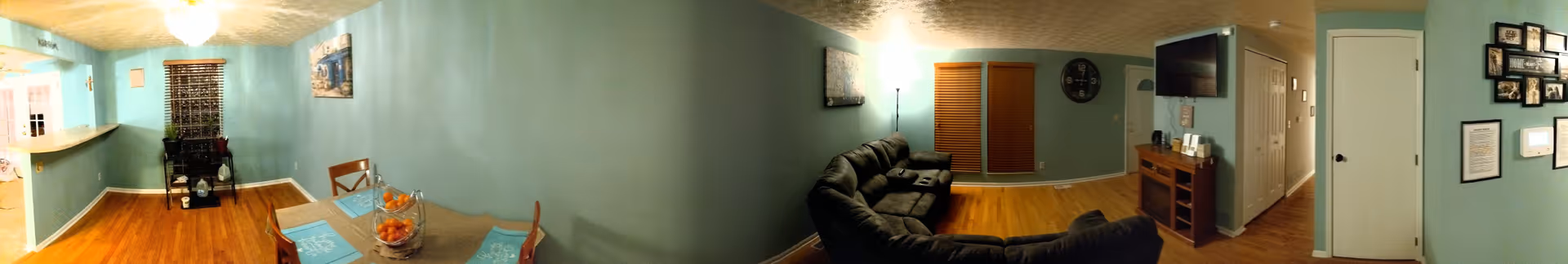 Panoramic view of a senior care home interior showing a dining area with a table set with placemats and a fruit basket, a living room with a dark sectional sofa, a wall-mounted TV, wooden blinds on the windows, light blue walls, hardwood floors, and various wall decorations including a clock and framed pictures.