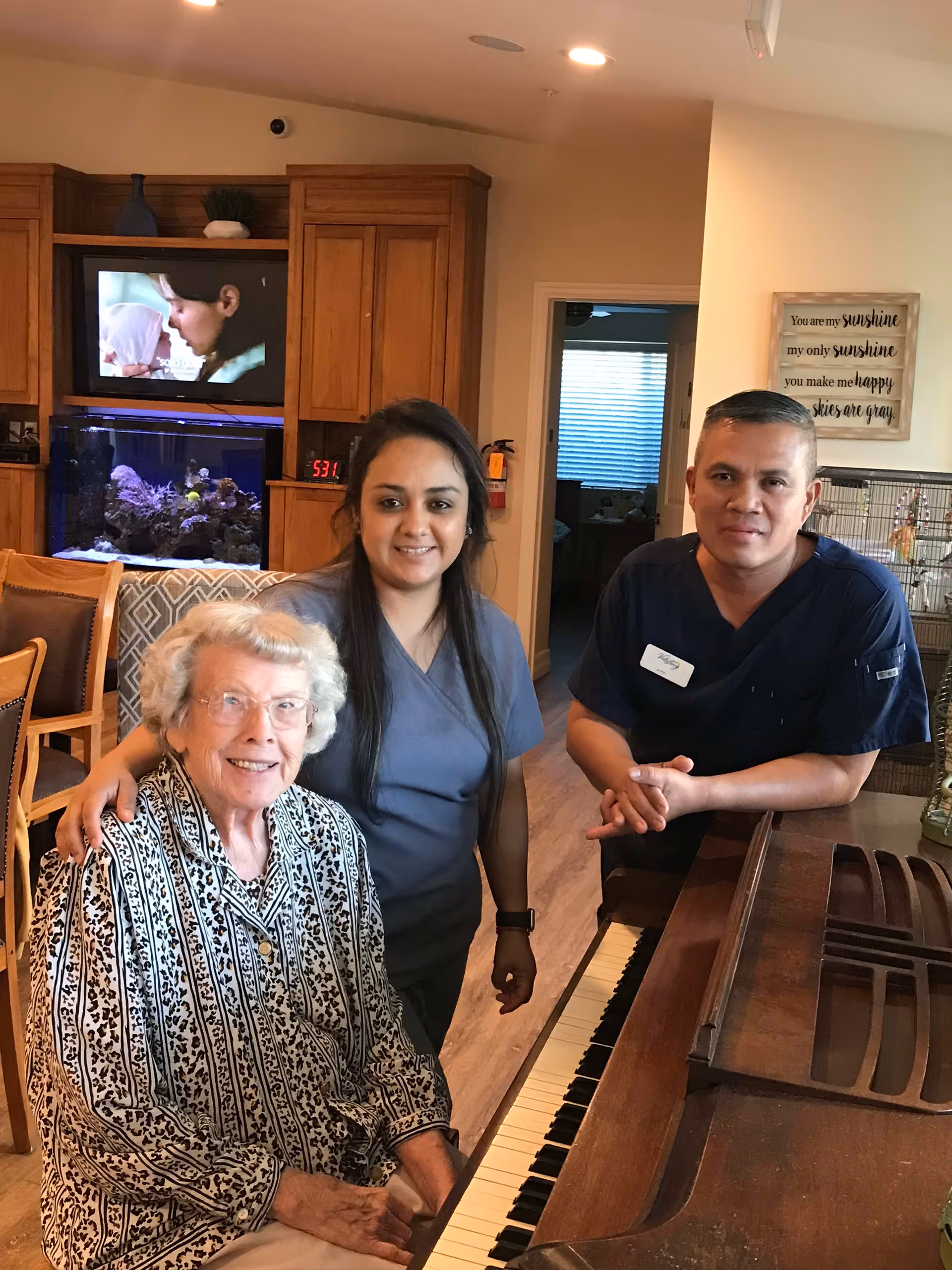 An elderly woman sitting at a piano with two caregivers standing beside her in a cozy living room area. Behind them is a wooden cabinet with a television and an aquarium, and a wall decoration with an inspirational quote.