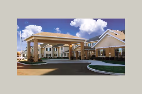 Exterior view of Winterberry Heights Senior Living community building with a covered entrance, beige walls, multiple windows, and a clear blue sky with clouds.