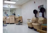 Interior view of a senior living facility's hair salon area with beige salon chairs, hair dryers mounted on the wall, a counter with a mirror, and a clock on the wall.
