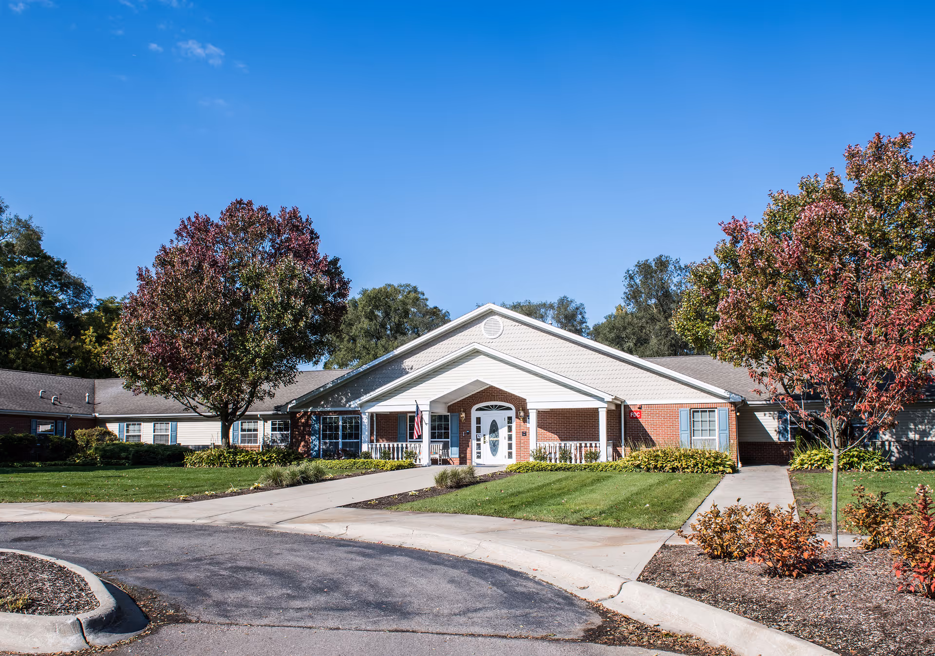 Front exterior view of Arden Courts - ProMedica Memory Care Community in Livonia, showing a single-story building with a white entrance porch, brick and siding facade, surrounded by green lawns and trees with autumn foliage under a clear blue sky.