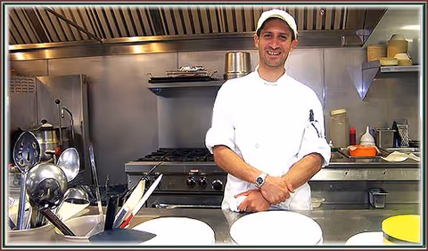 A chef wearing a white uniform and cap stands behind a stainless steel counter in a commercial kitchen, smiling at the camera. Various kitchen utensils and cooking equipment are visible around him.