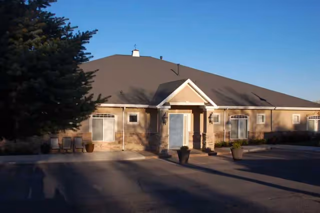 Exterior view of a single-story building with a pitched roof, beige walls, and stone accents. There are several windows and a central entrance door with a small covered porch. Two large potted plants are placed near the entrance, and a large tree is visible on the left side. The sky is clear and blue.