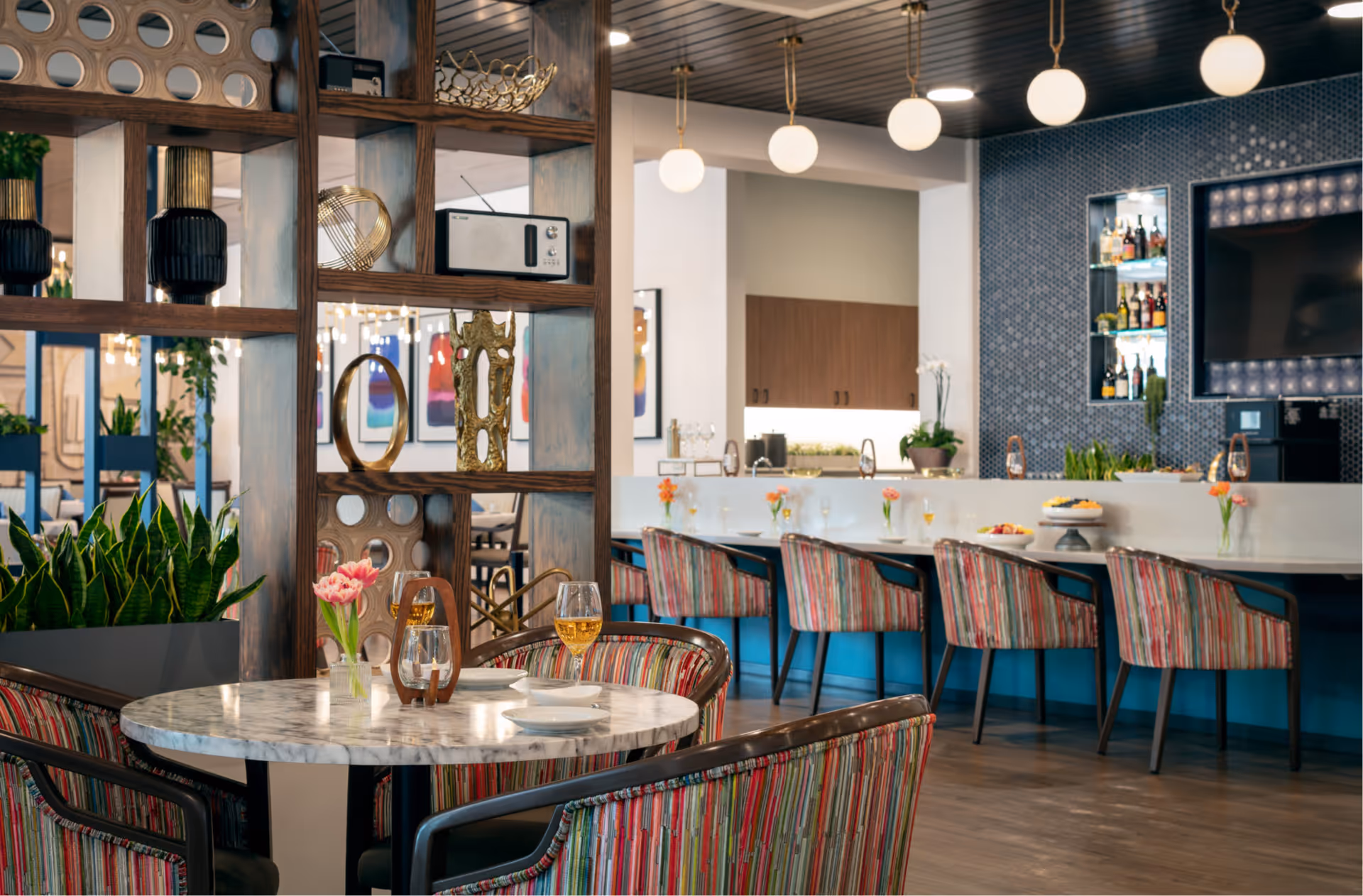 A modern dining area with a round marble table set with plates, glasses, and a small vase with pink flowers. Surrounding the table are colorful striped chairs. In the background, there is a bar counter with more striped chairs, small flower vases, and a display of bottles on a blue tiled wall. The space is decorated with hanging globe lights and wooden shelving with decorative items.