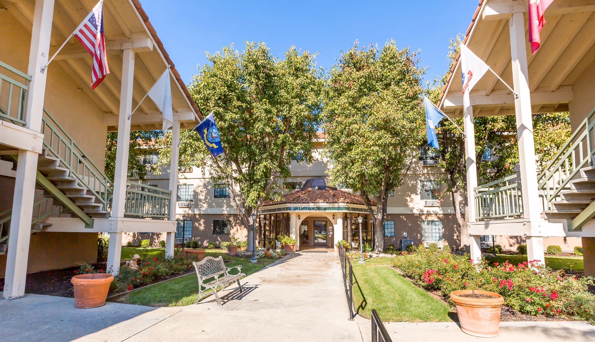 Exterior view of Brookdale Tracy senior living facility entrance with a pathway leading to the main door, flanked by green lawns, flower beds, benches, and several flags mounted on the building. The building has a beige facade with a tiled roof and multiple windows.