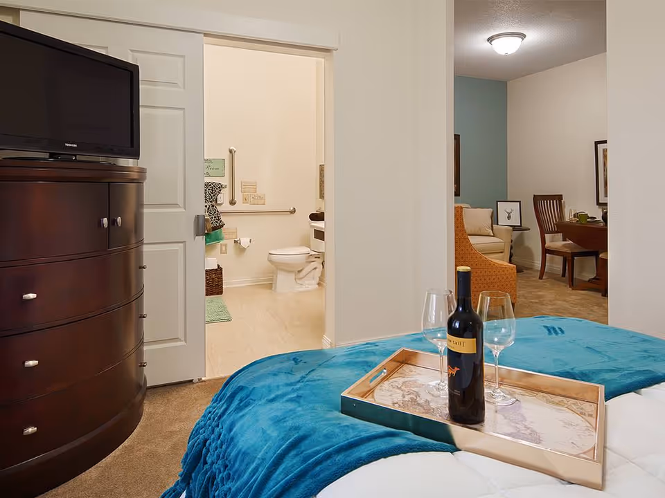 View of a bedroom with a teal blanket and a tray holding a bottle of wine and two wine glasses on the bed. To the left, there is a dark wooden dresser with a TV on top. In the background, an open door reveals a bathroom with a toilet and grab bars. To the right, there is a living area with a sofa, an orange armchair, and a dining table with chairs.
