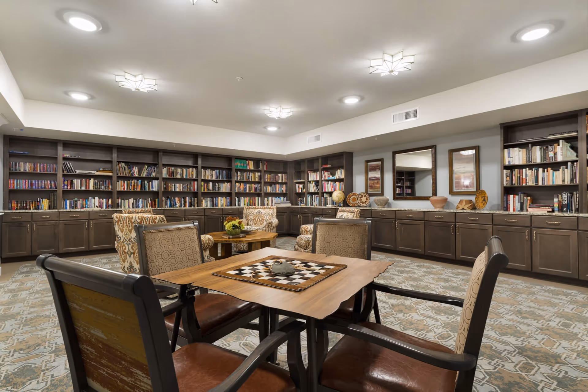A cozy senior living community library and lounge area with bookshelves filled with books along the walls, comfortable upholstered chairs, a wooden table with a checkerboard game, decorative pottery, and framed artwork on the walls.