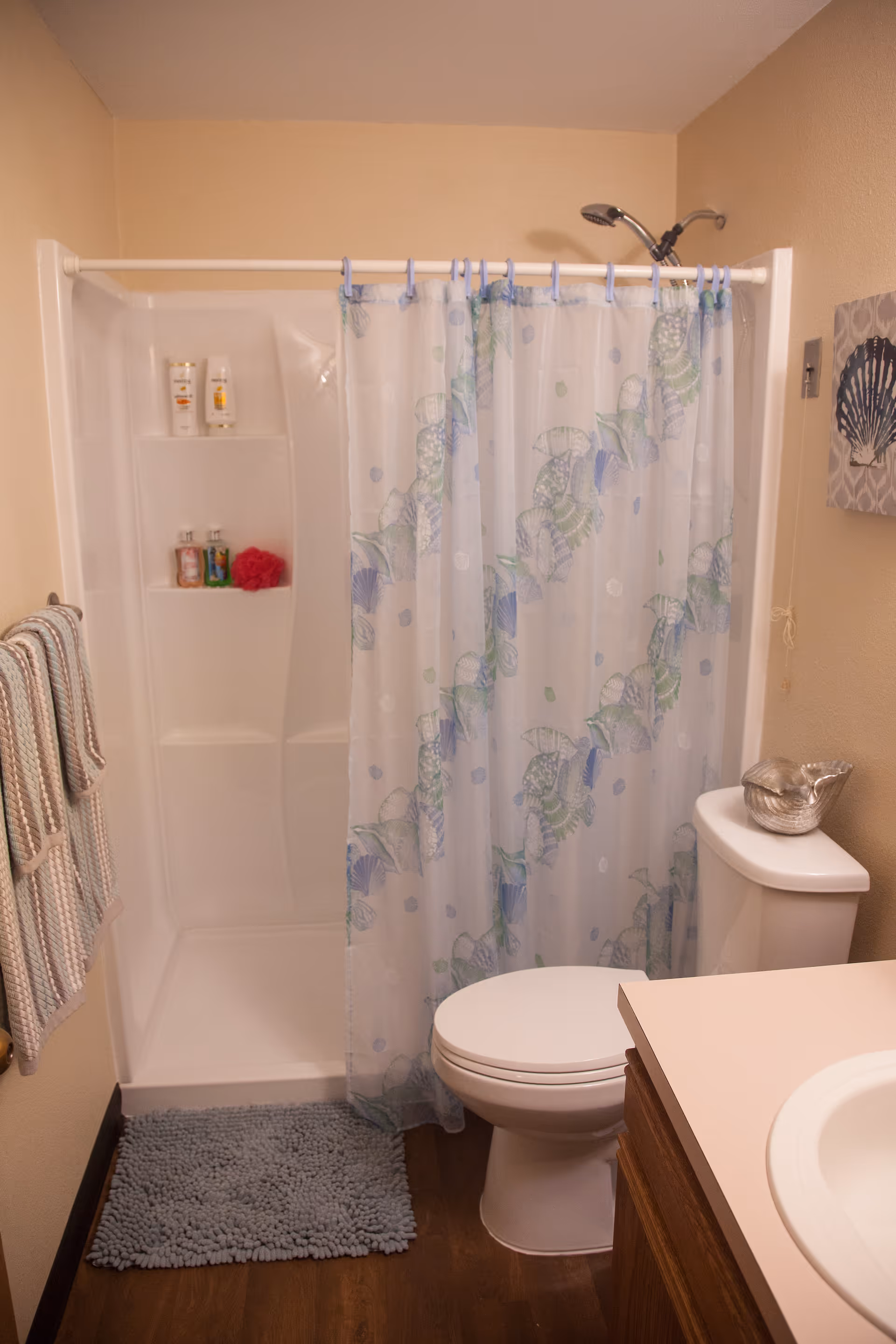 A small bathroom featuring a white shower with built-in shelves holding toiletries, a shower curtain with a seashell pattern, a white toilet with a decorative shell on the tank, a wooden vanity with a white countertop and sink, a blue bath mat on the wooden floor, and striped towels hanging on a rack on the wall.