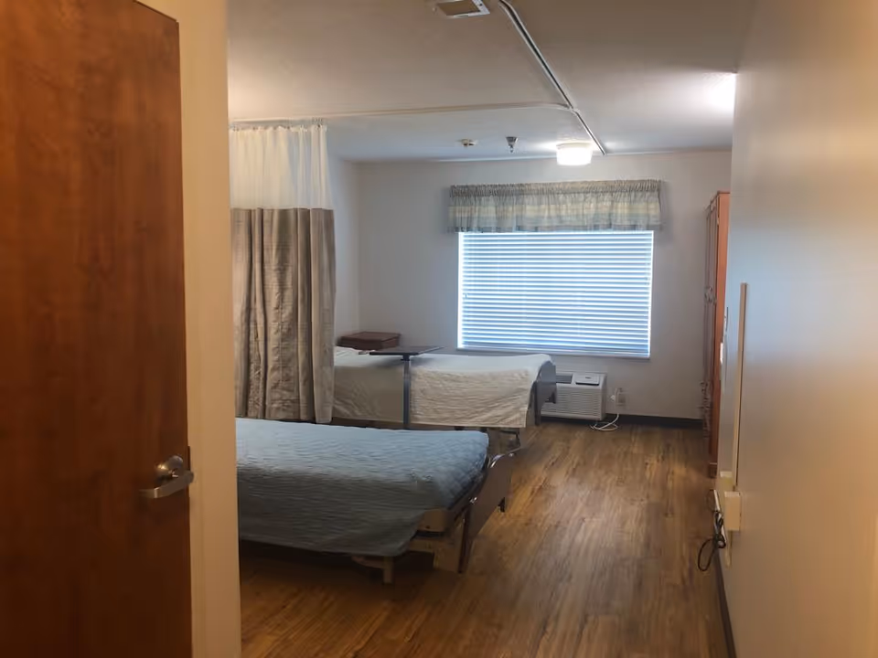 A room with two hospital-style beds separated by a curtain. Each bed has a light-colored bedspread, and there is a small overbed table on one bed. The room has wood flooring, a window with blinds and a valance, and a wooden cabinet on the right side.