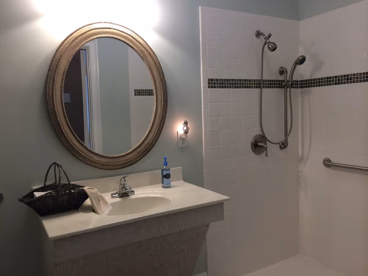 A bathroom with a white sink countertop, a round decorative mirror above the sink, a basket with a towel on the countertop, a soap dispenser, and a tiled shower area with a handheld showerhead and a grab bar.