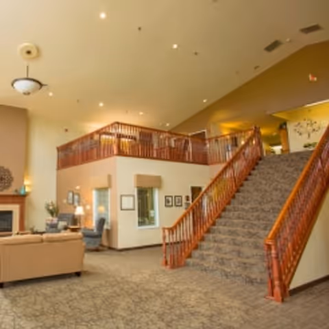 Interior view of a senior living facility featuring a carpeted staircase with wooden railings leading to an upper level. The area includes a seating space with sofas and armchairs near a fireplace, soft lighting, and framed pictures on the walls.