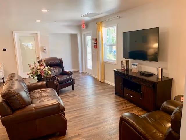 A cozy living room with brown leather sofas and armchairs arranged around a dark wooden TV stand with a flat-screen television mounted on the wall. The room has wood flooring, a window with yellow curtains, and a door leading outside. A vase with flowers is placed on a side table next to one of the armchairs.