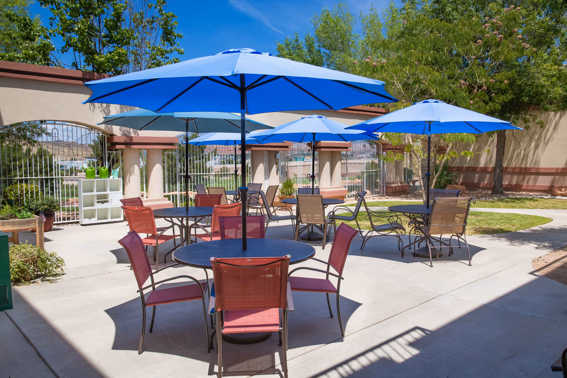 Outdoor patio area with several round tables, each shaded by large blue umbrellas. The tables are surrounded by red and brown chairs. The patio is enclosed by a beige wall with decorative arches and metal gates, with trees and greenery visible around the area under a clear blue sky.