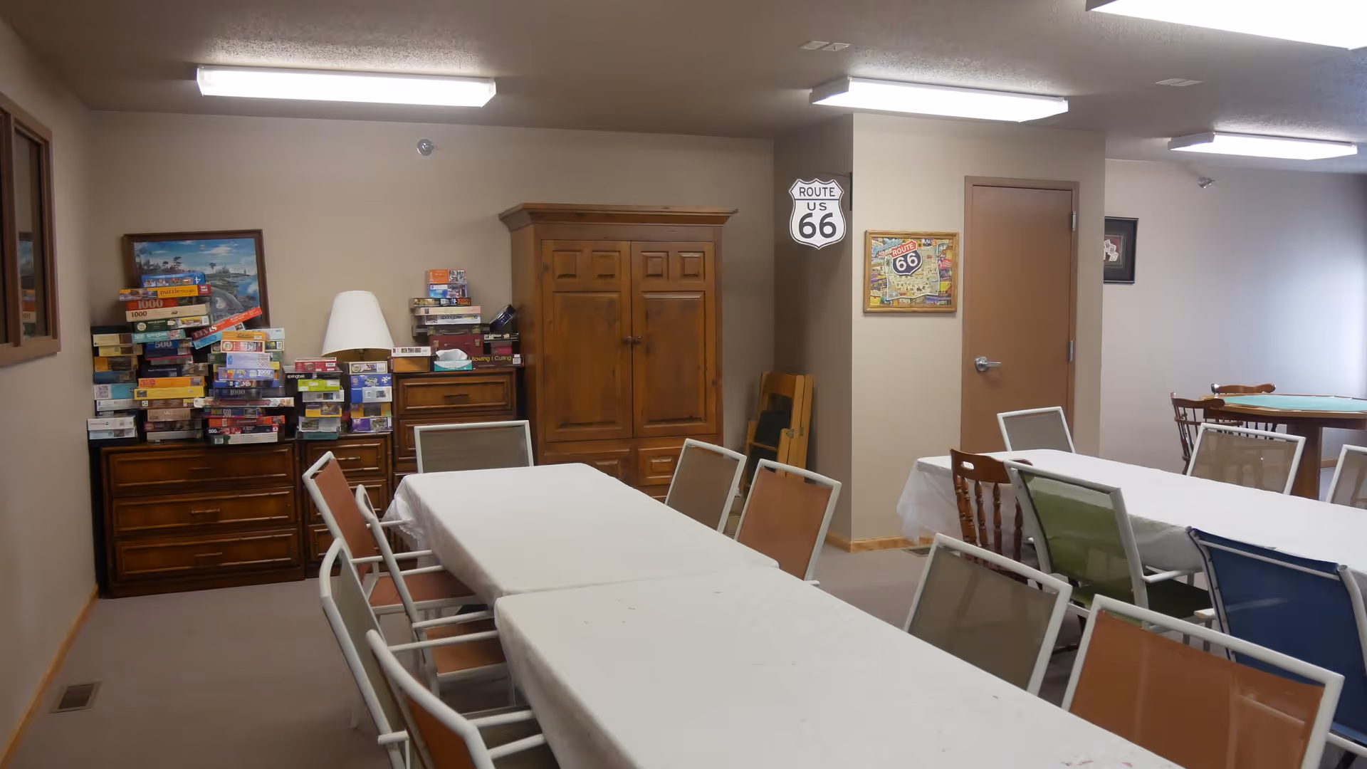 A community room with several tables covered in white tablecloths and surrounded by various colored chairs. Against the wall, there are wooden cabinets stacked with board games and puzzles. The walls are decorated with Route 66 signs and framed pictures. The room is lit by fluorescent ceiling lights.