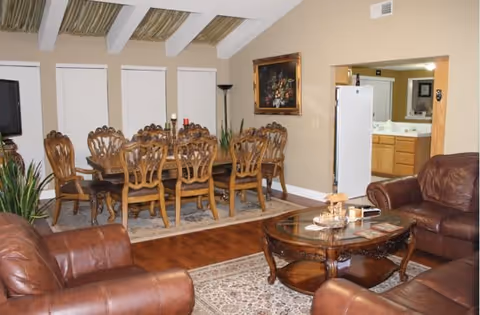 A cozy living and dining area featuring brown leather sofas arranged around a glass coffee table with decorative items. The dining area has a wooden table with eight ornate chairs on a rug. The room has wooden flooring, beige walls, a painting on the wall, and a view into a kitchen area with a white refrigerator and wooden cabinets.
