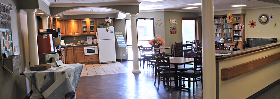 Interior view of a senior living facility common area featuring a small kitchen with wooden cabinets, a white refrigerator, and a microwave. Adjacent to the kitchen are several dining tables with chairs, a bulletin board on the left wall, and a bookshelf with books and decorations on the right. The space is well-lit with ceiling lights and natural light from a door and windows.