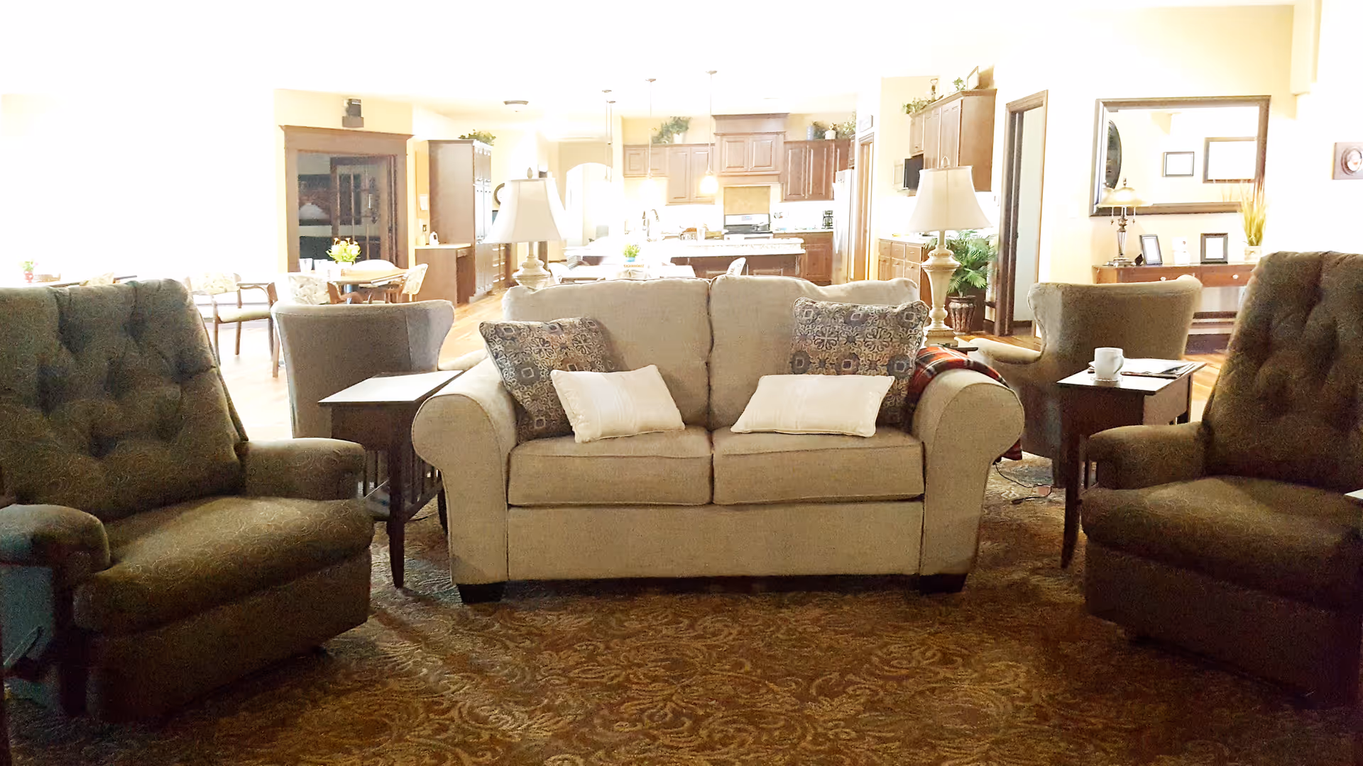 Comfortable common-area living room with a beige sofa flanked by two armchairs, side tables and lamps, opening onto a dining and kitchen area.