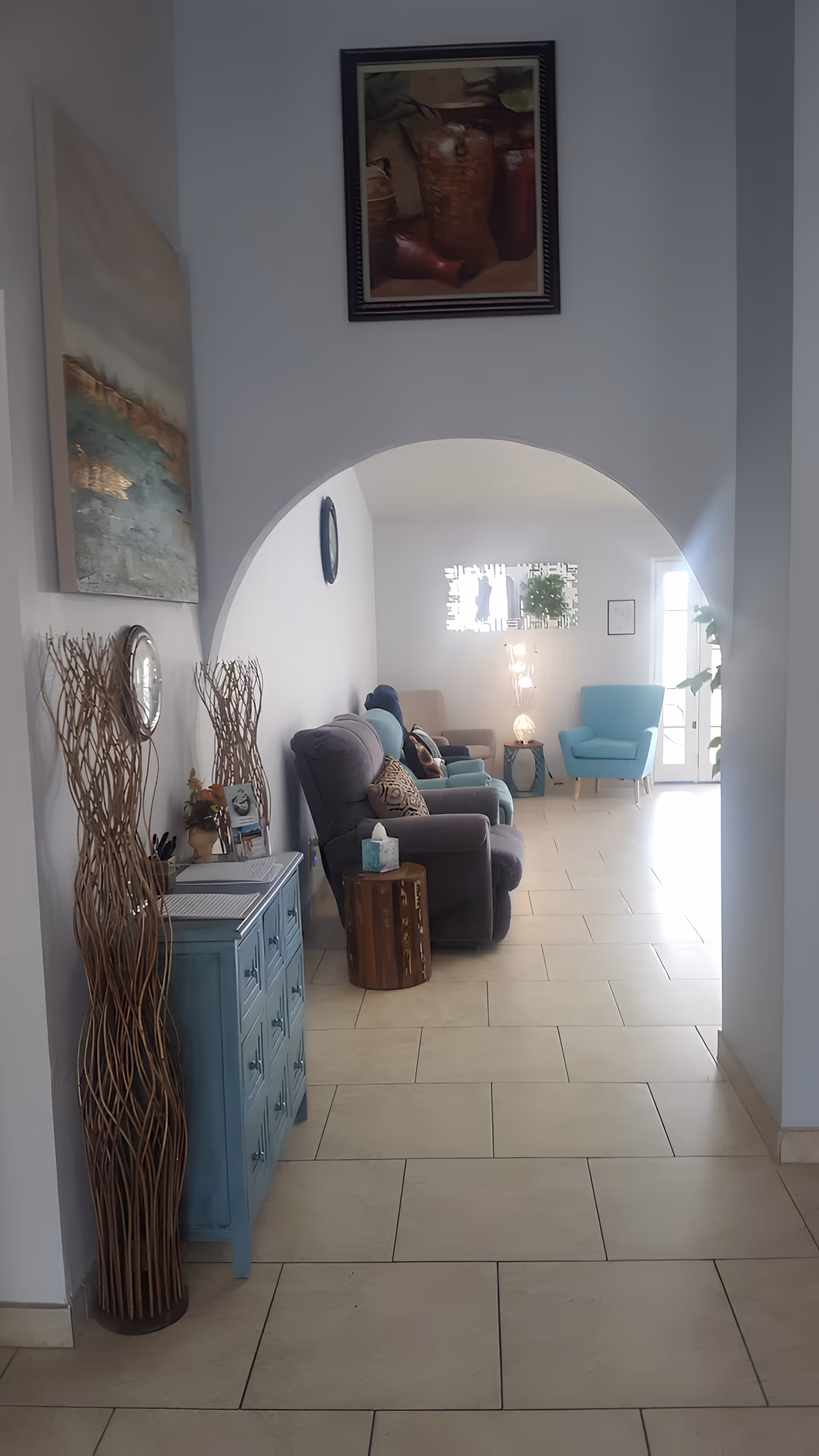 A bright and cozy living room area in Living Waters Care Home featuring a row of comfortable armchairs in various shades of blue and gray. The room has light-colored tiled flooring, white walls, and an arched doorway. Decorative elements include a blue chest of drawers, tall wicker vases, framed artwork on the walls, a small wooden side table with a tissue box, and a modern lamp on a side table near the window.