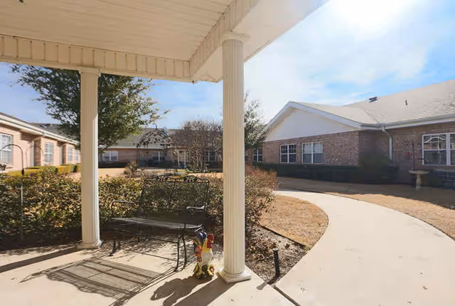 Covered patio with a bench and small decorative statue facing a courtyard and single-story brick senior living buildings.