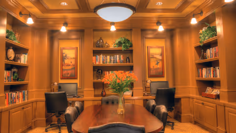 A warm, well-lit office or study room with a wooden table and six black leather chairs around it. The room has built-in wooden bookshelves filled with books and decorative items, two computer workstations with black office chairs, framed landscape paintings on the walls, and green plants placed on top of the shelves. The lighting includes wall sconces and a central ceiling light fixture.