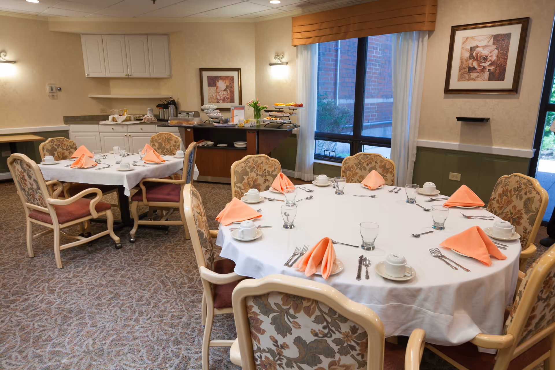 A dining room with two round tables covered with white tablecloths, each set with cups, glasses, silverware, and peach-colored folded napkins. The room has floral upholstered chairs, a carpeted floor, a window with curtains, and a buffet area with plates and food items in the background.