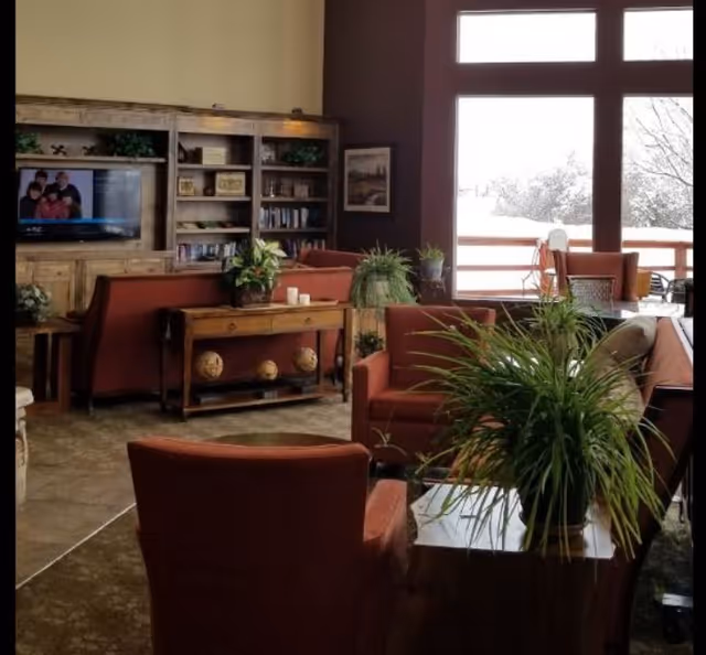 A cozy living room area with red upholstered chairs and sofas arranged around a wooden coffee table with green plants. There is a wooden entertainment center with a TV and bookshelves against the wall. Large windows in the background show a snowy outdoor scene.