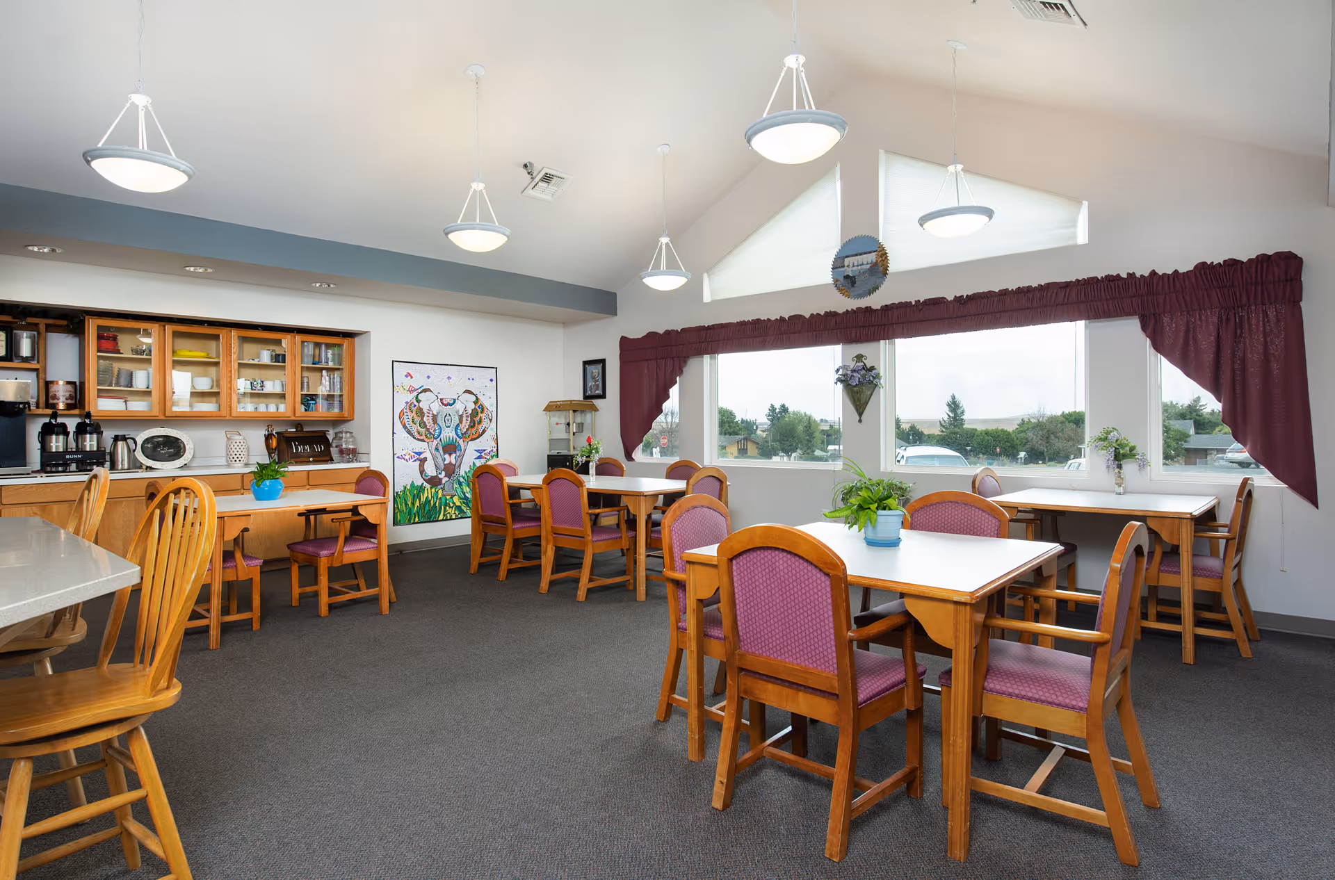 A bright dining room with several wooden tables and chairs with purple cushions. The room has large windows with maroon curtains, letting in natural light and offering a view of trees and houses outside. There are hanging ceiling lights and a cabinet with glass doors displaying dishes and kitchen items. A colorful painting of an animal is on one wall, and there are small plants on the tables and windowsills.
