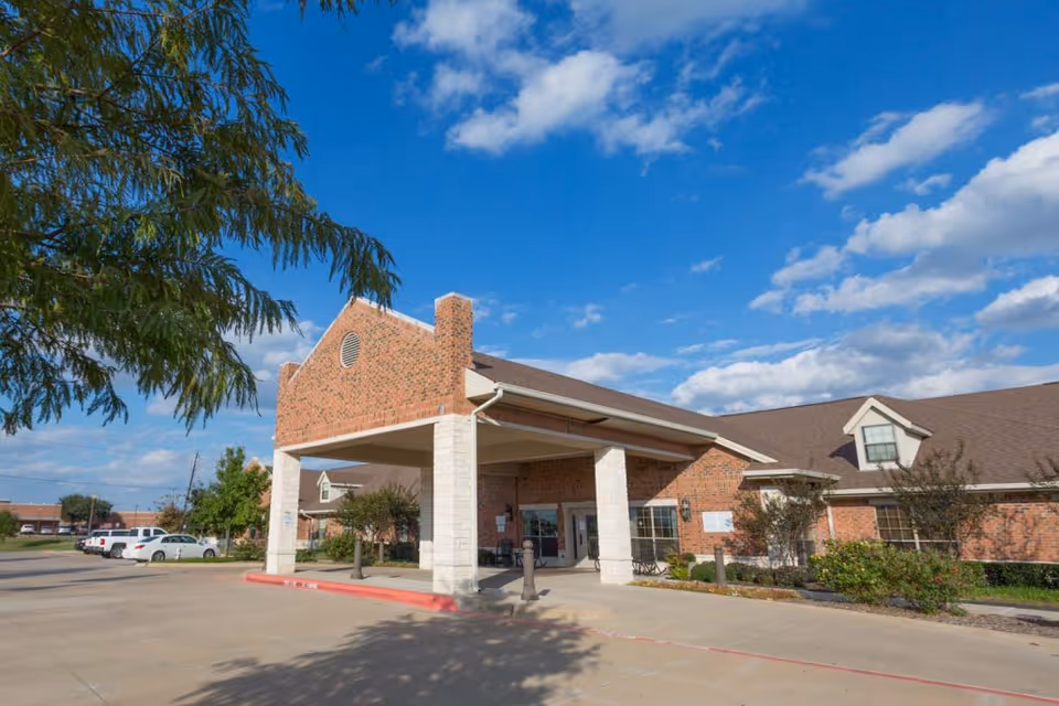 Exterior view of a brick building with a covered entrance and a parking lot under a blue sky with scattered clouds. The building has a pitched roof with dormer windows and some landscaping with bushes and small trees near the entrance.