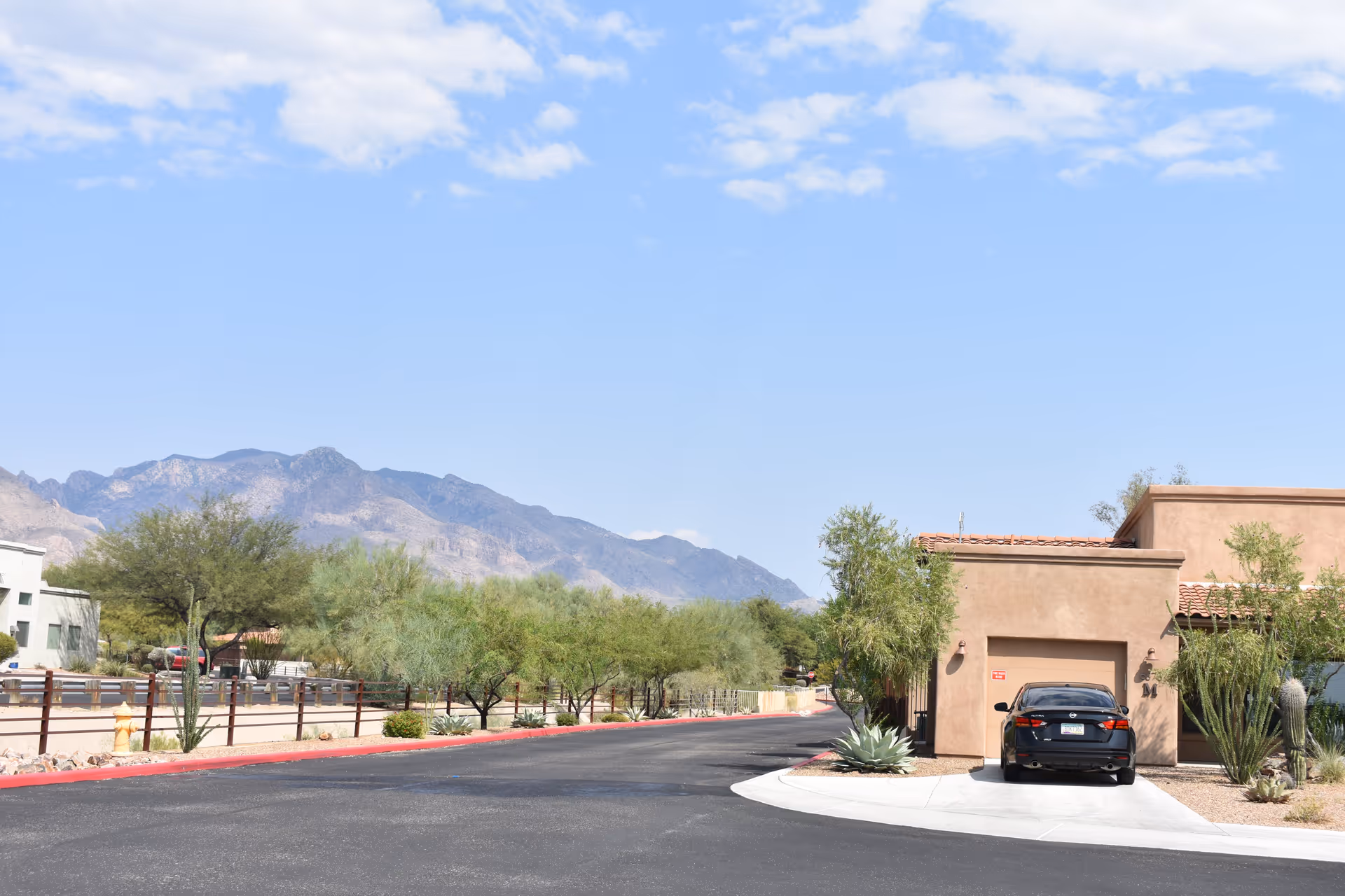 A paved road in a senior living community with a single-story building on the right featuring a garage and a parked black car. Desert landscaping with trees and shrubs lines the road, and mountains are visible in the background under a partly cloudy blue sky.