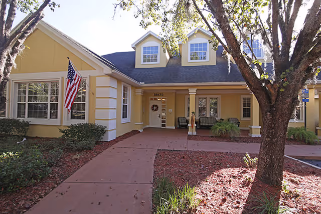 Front entrance of a yellow senior living building with a covered porch, American flag, and trees along the walkway.