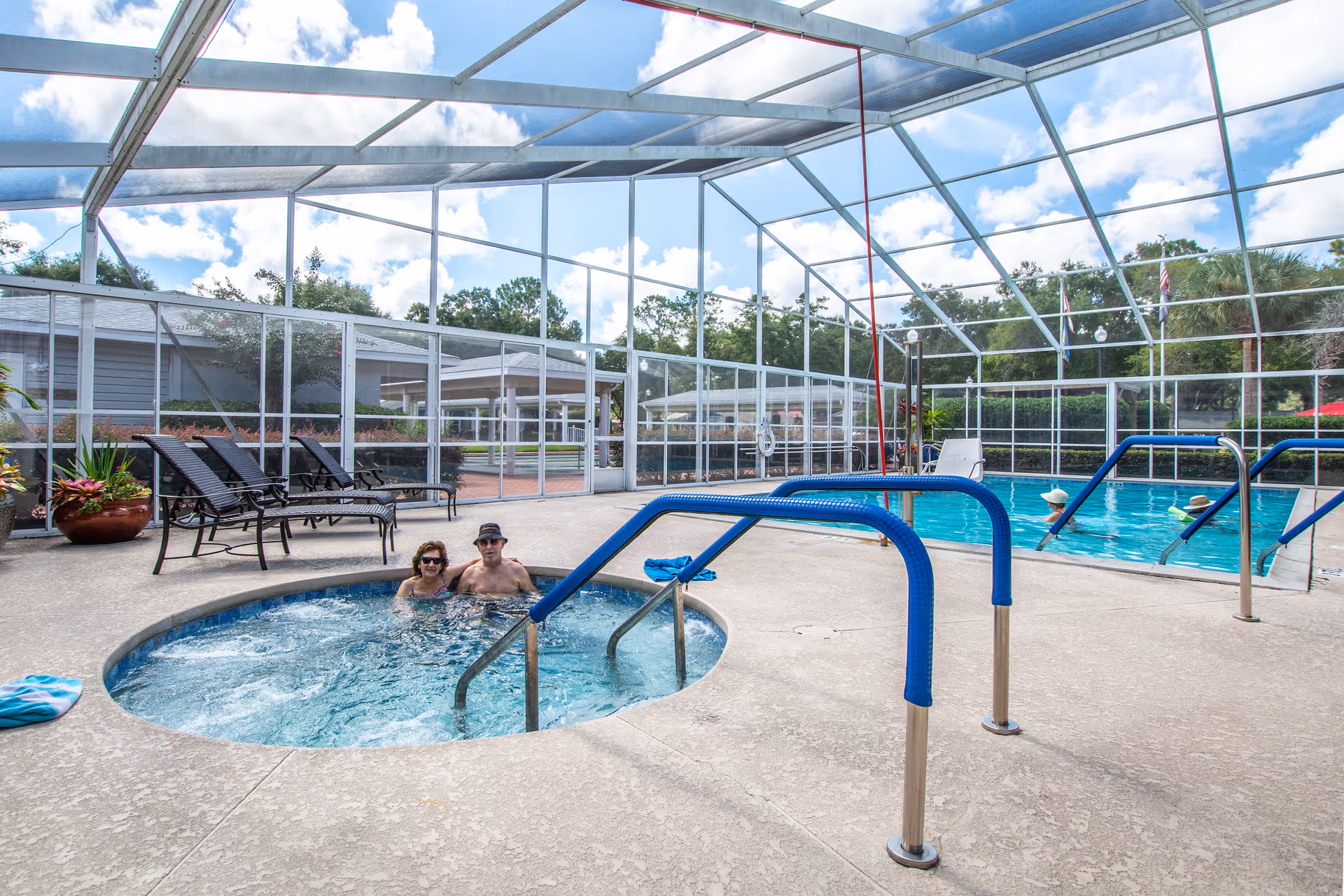 Two people relaxing in a circular hot tub next to a swimming pool inside a screened enclosure with lounge chairs and plants nearby under a partly cloudy sky.