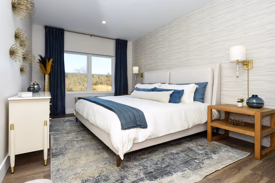 Well-lit modern bedroom with a large upholstered bed dressed in white and blue linens, nightstands, decorative wall art, and a window with dark curtains.